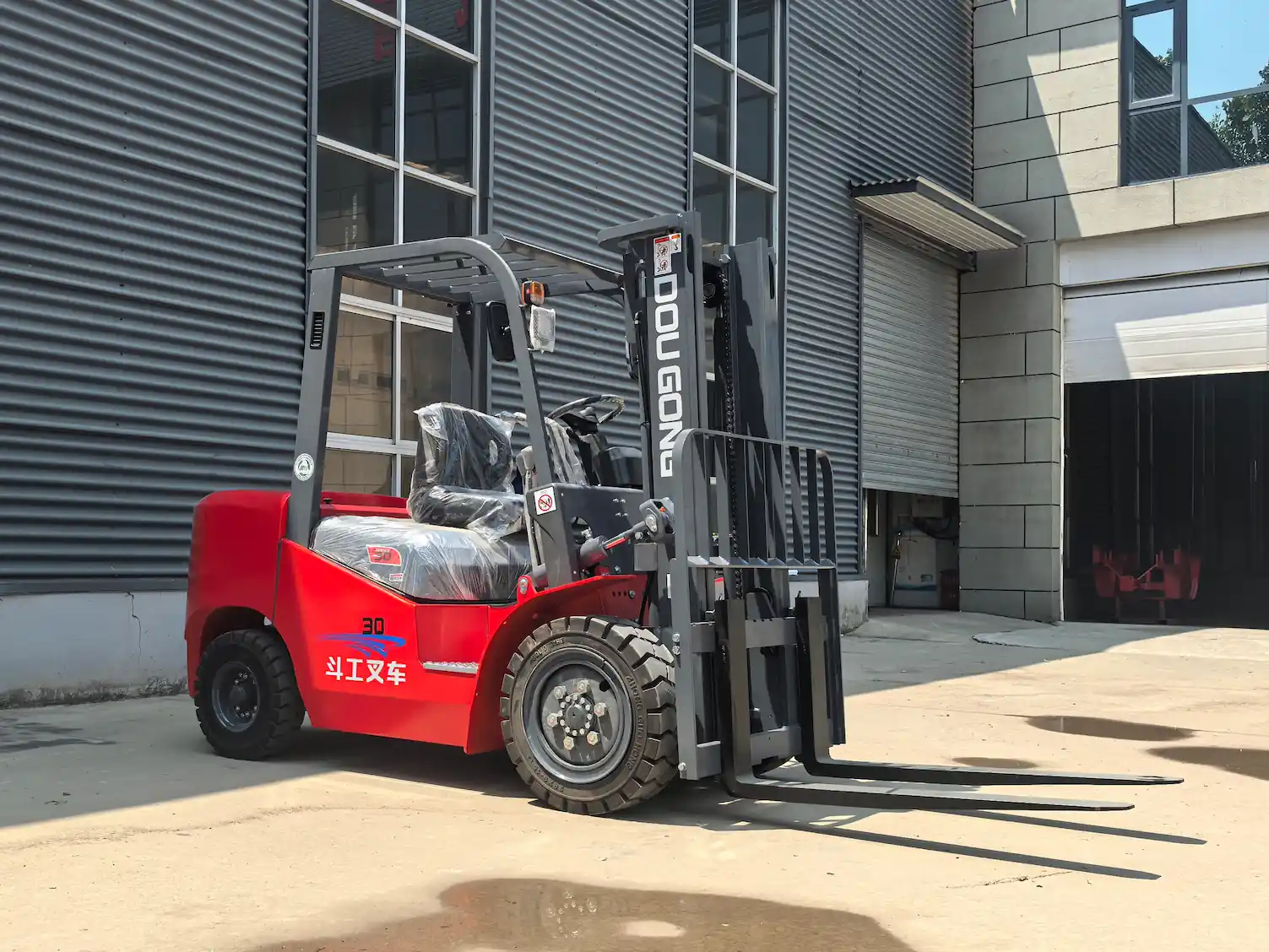 Red 3-ton DOGON forklift parked outdoors in front of an industrial building, showing forks, tires, and protective seat covers.