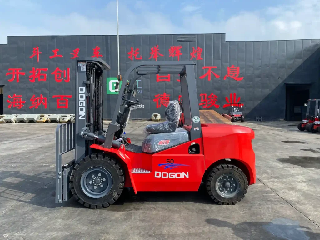Red DOGON 5-ton diesel forklift parked outside a warehouse on a concrete surface, with protective plastic covering the seat, clear view of tires and lifting mechanism, and a gray industrial building with red Chinese characters in the background.